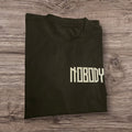 T-shirt nera in cotone Ottoman Kumus con scritta Nobody bianca sul petto, stile streetwear piegata su legno