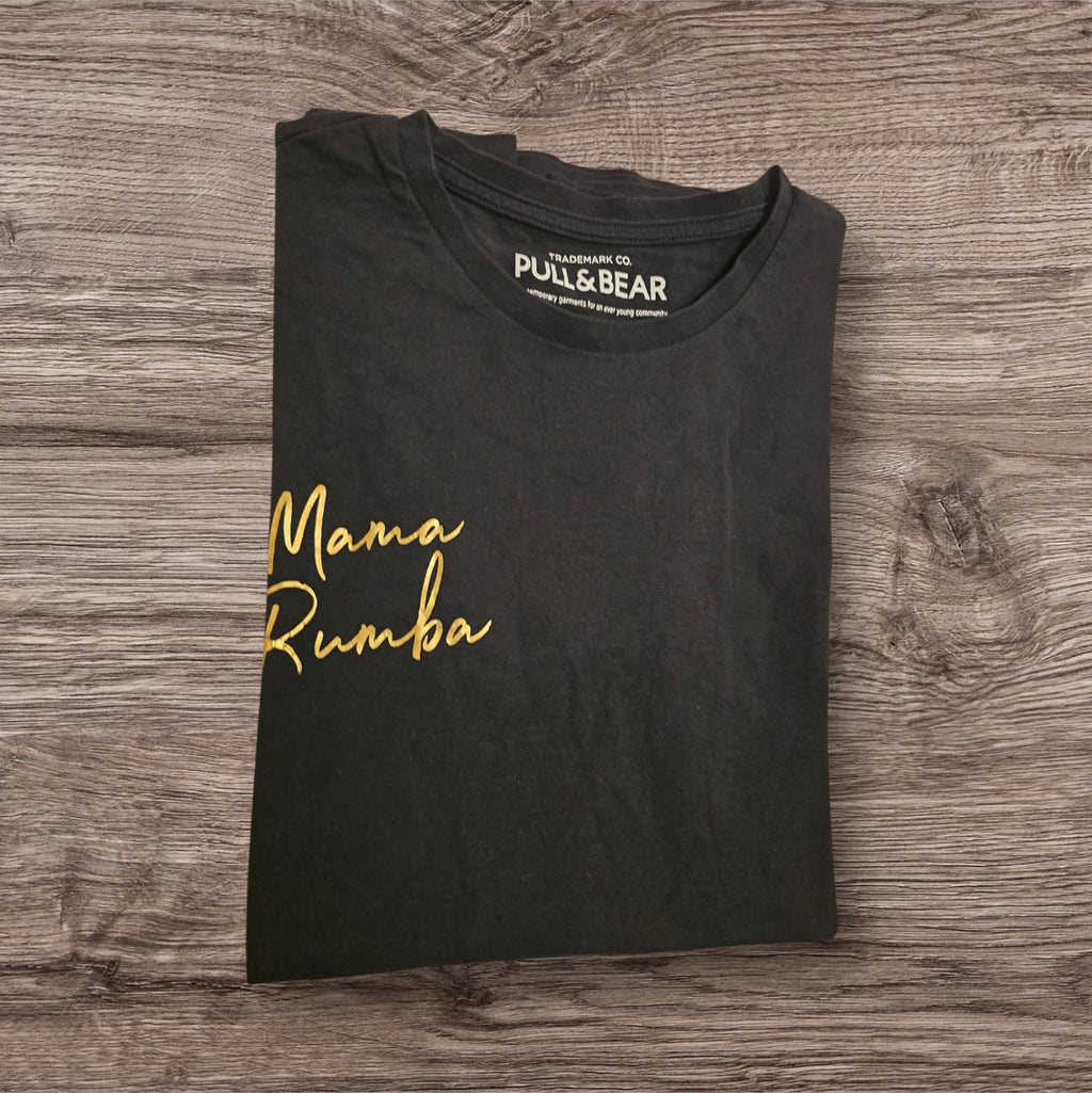 T-shirt nera Ottoman Kumus PULL&BEAR in cotone con scritta Mama Rumba dorata sul petto, piegata su sfondo in legno.