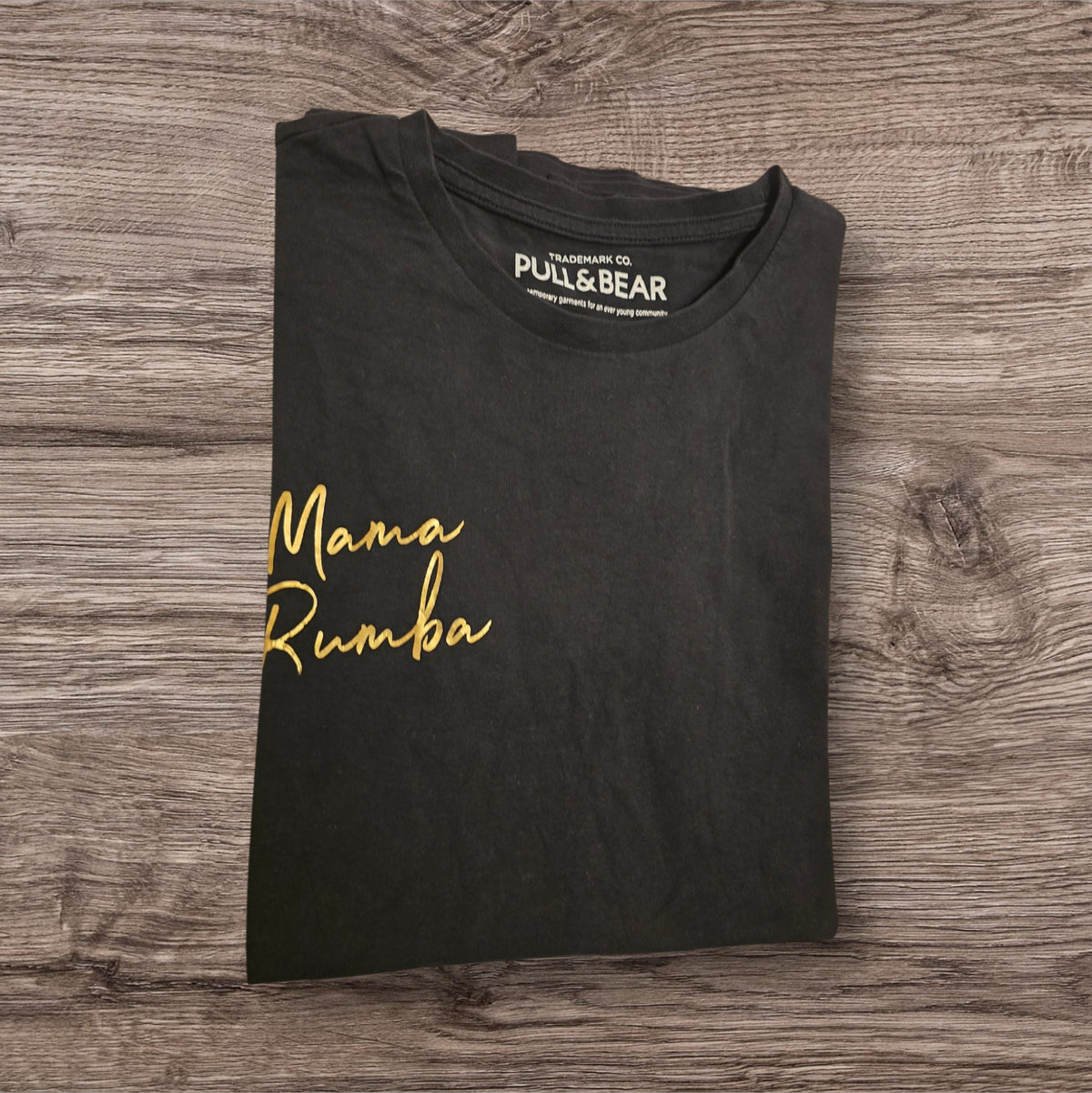 T-shirt nera Ottoman Kumus PULL&BEAR in cotone con scritta Mama Rumba dorata sul petto, piegata su sfondo in legno.