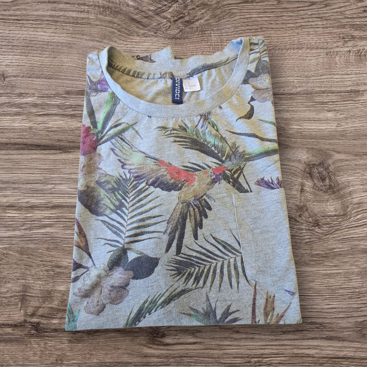 T-shirt Ottoman Kumus grigio mélange con stampa tropicale di pappagalli e taschino sul petto, piegata su legno.